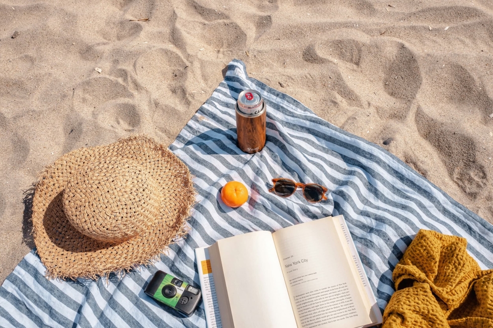 Een handdoek die in het zand ligt met daarop een heleboel zomerspullen zoals een zonnebril, zonnehoed en frisdrank
