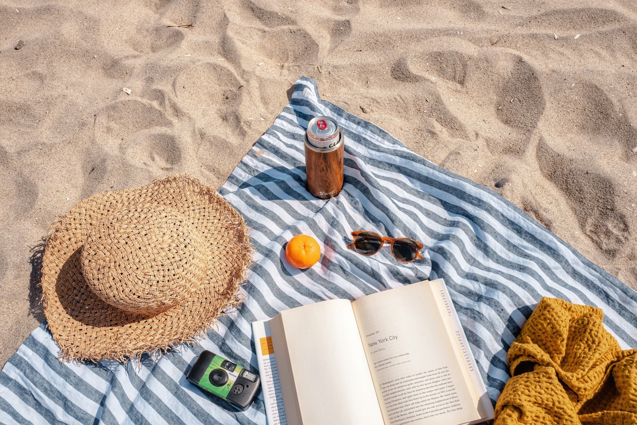 Een handdoek die in het zand ligt met daarop een heleboel zomerspullen zoals een zonnebril, zonnehoed en frisdrank