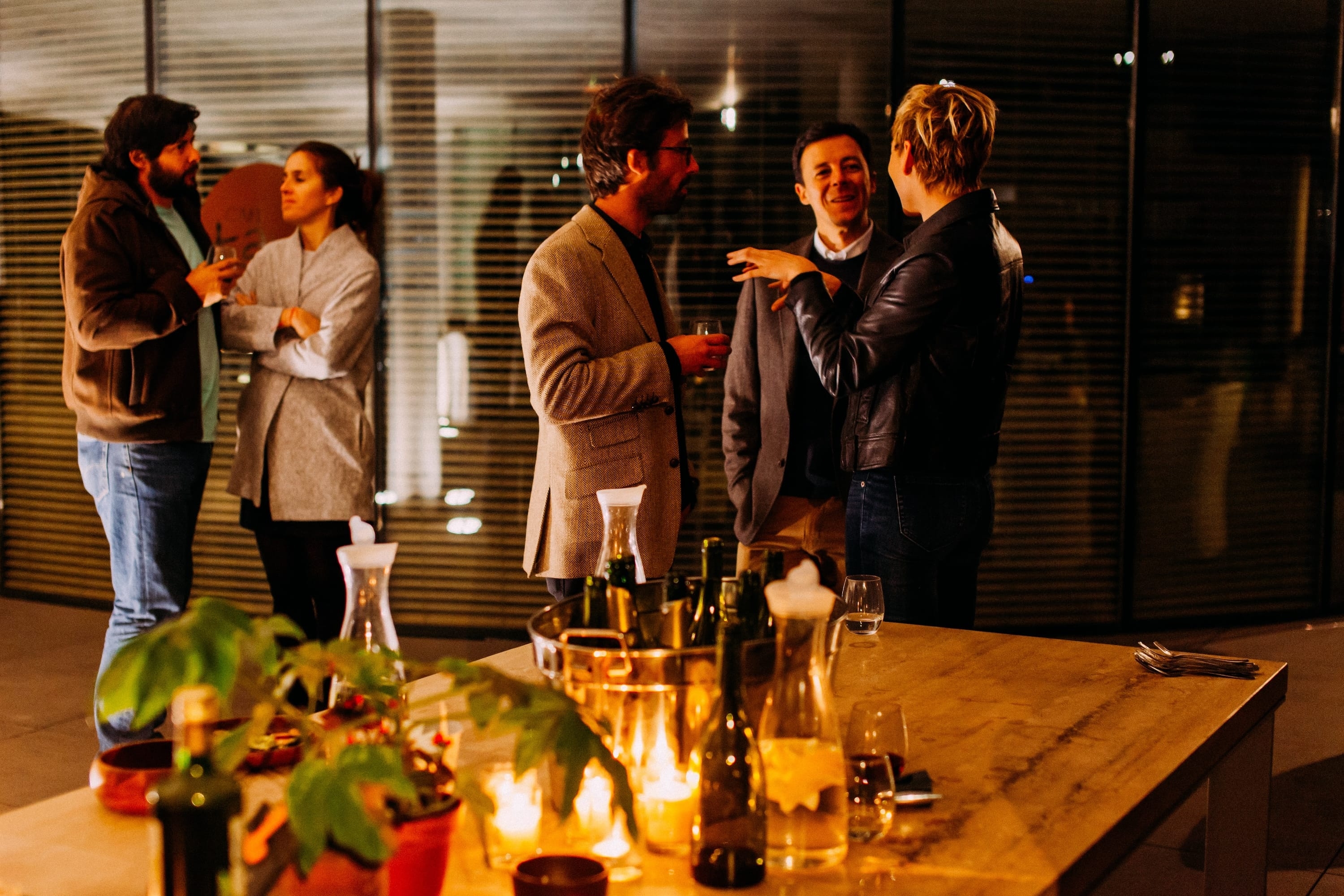 Een warme sociale bijeenkomst met vijf personen in gesprek rond een houten tafel met kaarsen, glazen en planten