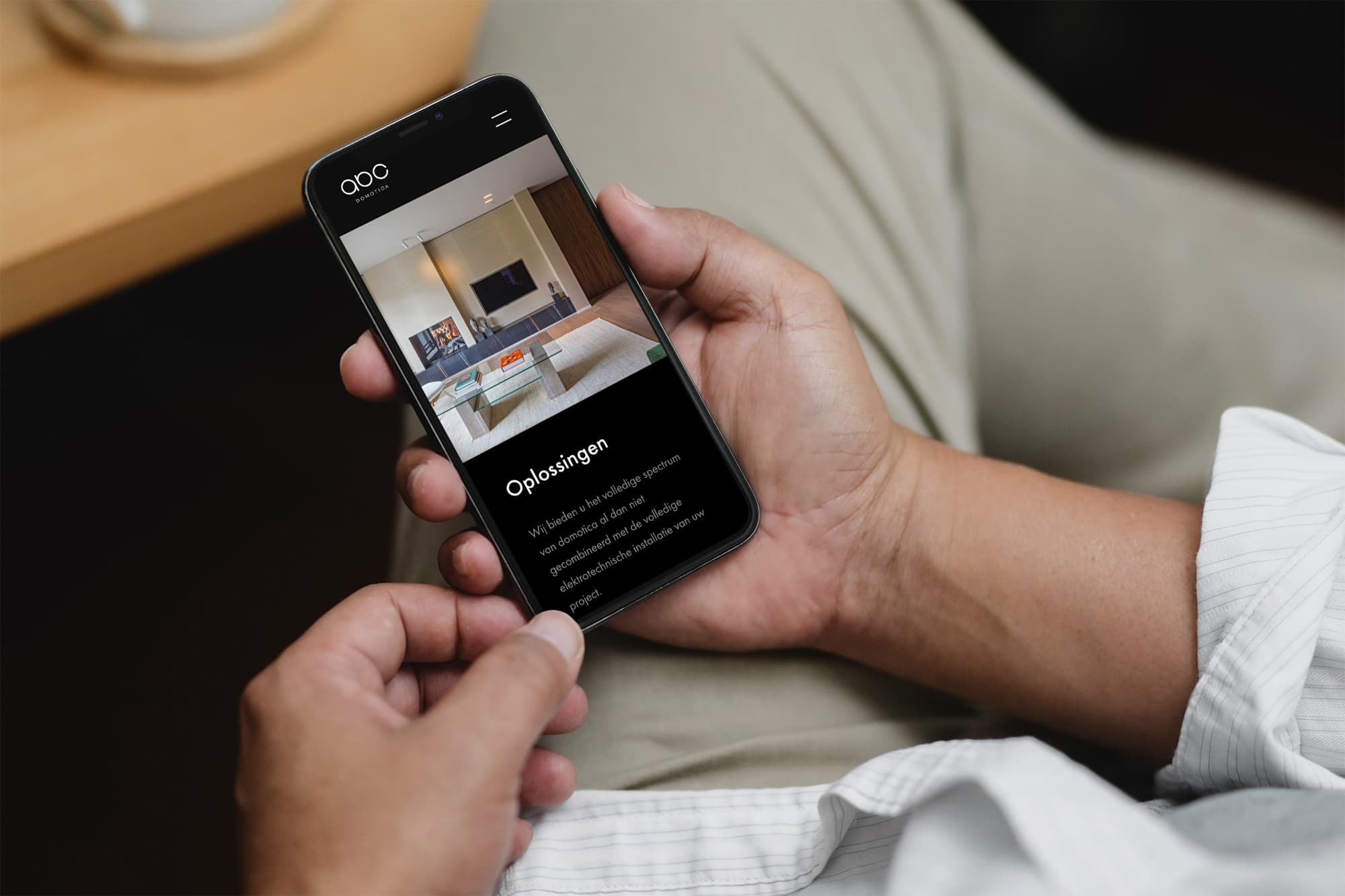 Een persoon die een smartphone vasthoudt met een website over domotica op het scherm, toont een moderne woonkamer met televisie en open haard