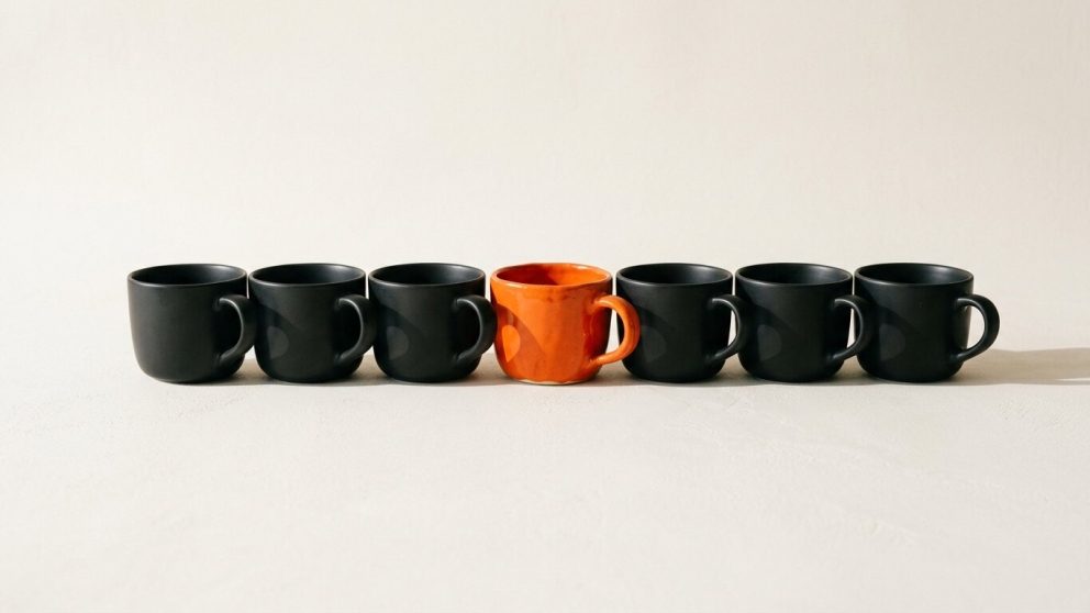 Een rij identieke zwarte kopjes met één handgemaakte, onregelmatige oranje kop ertussen, op een gebroken witte achtergrond