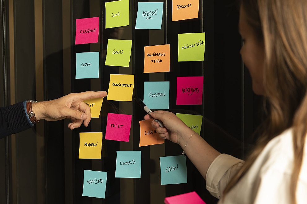 Maarten en een teamlid brainstormen over een webdesignstrategie in Antwerpen, waarbij ze kleurrijke sticky notes op een glazen wand gebruiken om ideeën te ordenen.