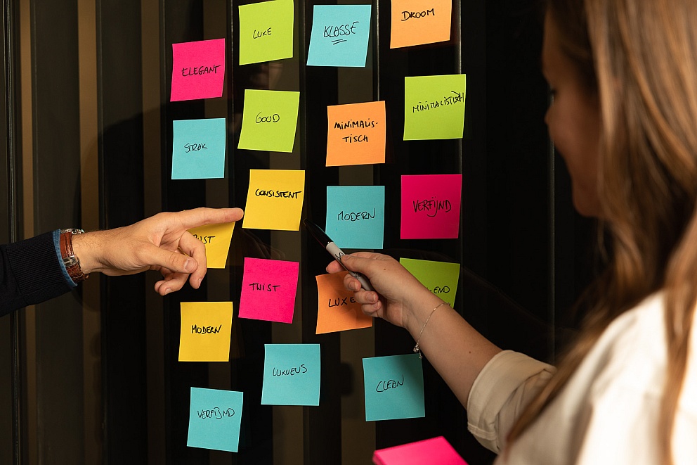 Maarten en een teamlid brainstormen over een webdesignstrategie in Antwerpen, waarbij ze kleurrijke sticky notes op een glazen wand gebruiken om ideeën te ordenen.